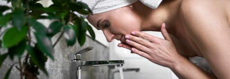 Femme se lavant le visage à l'eau du robinet dans une salle de bain lumineuse